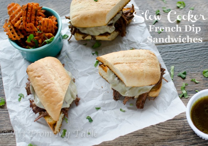 Three french dip sandwiches on a piece of parchment paper, served with au jus and sweet potato waffle fries.