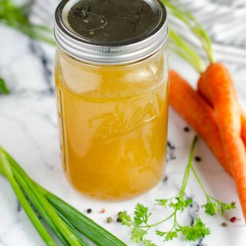 quart size mason jar filled with chicken bone broth, surrounded by carrots, celery, parsley and peppercorns