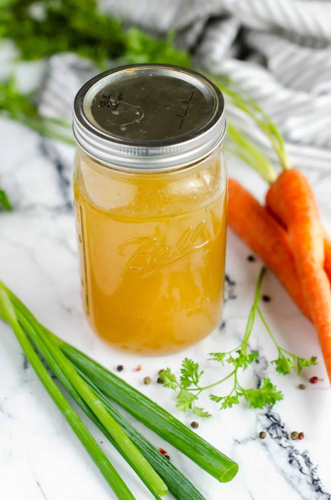 quart size mason jar filled with chicken bone broth, surrounded by carrots, celery, parsley and peppercorns