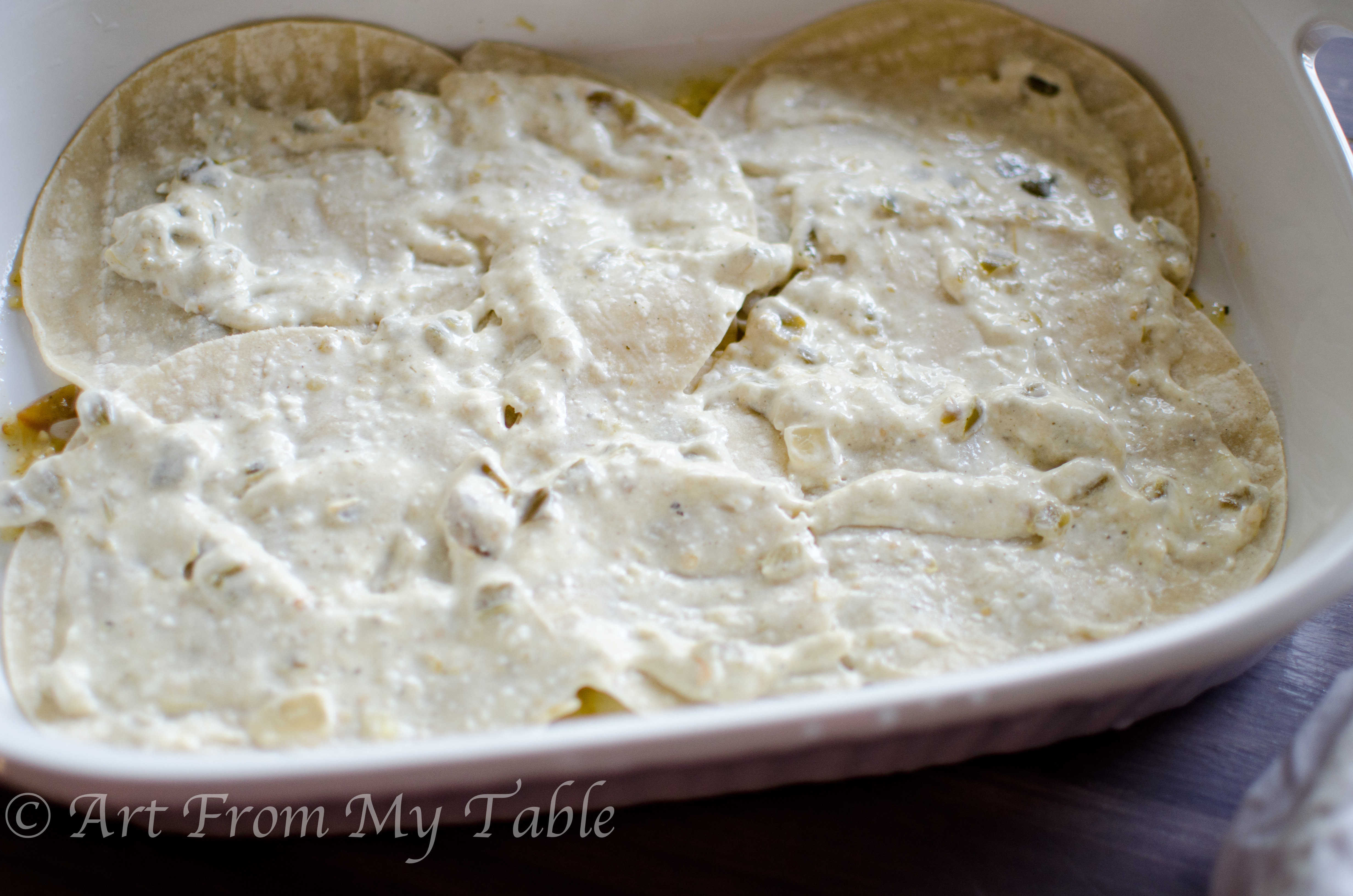 White casserole dish with a layer of tortillas and verde sauce.