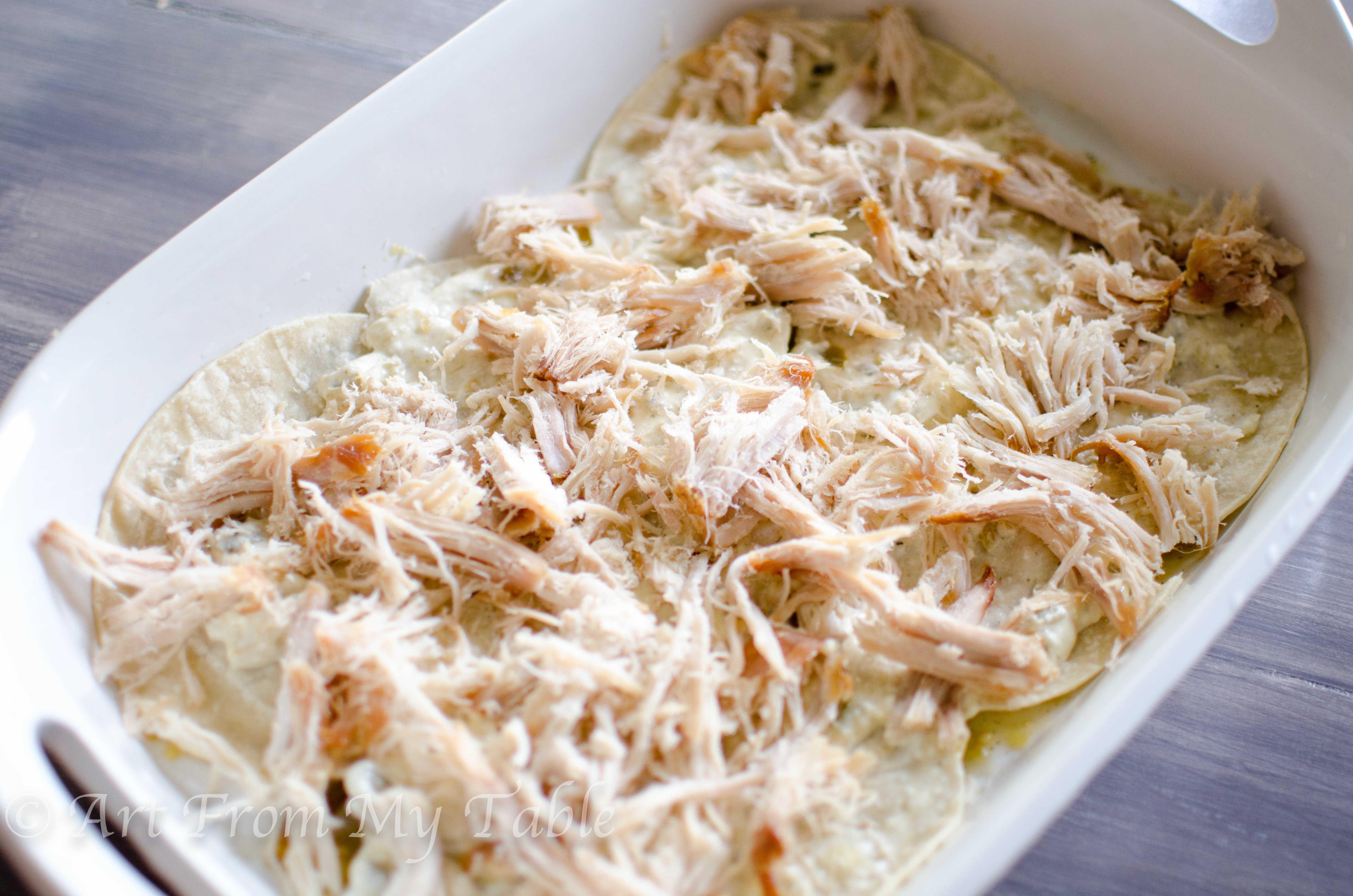 White casserole dish with layer of tortillas, verde sauce, and shredded pork.