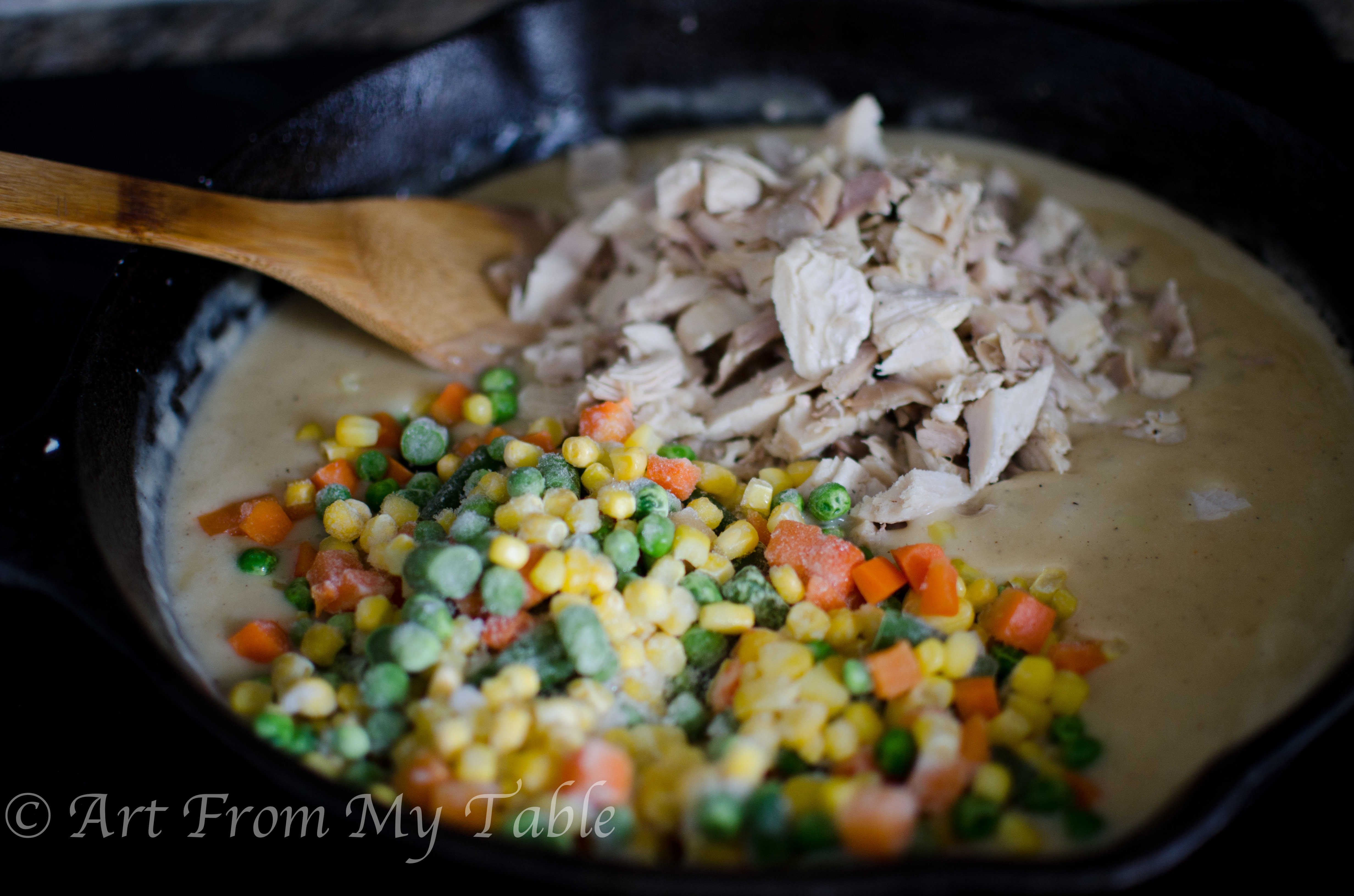 Cast iron skillet with wood spoon stirring the turkey and vegetables into the sauce of the turkey pot pie.