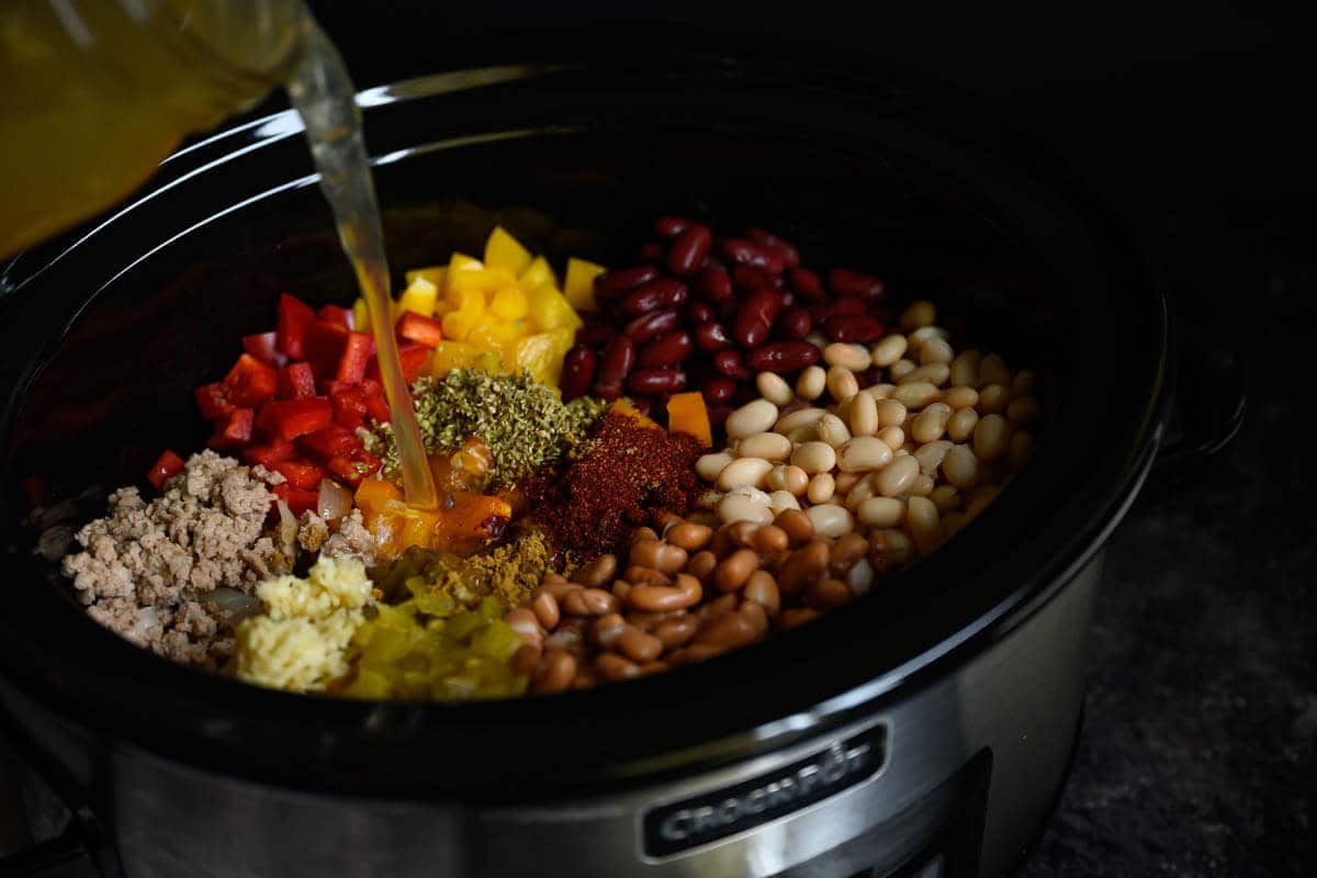 ground turkey, beans, peppers, and spices in a crockpot chicken broth being poured in.