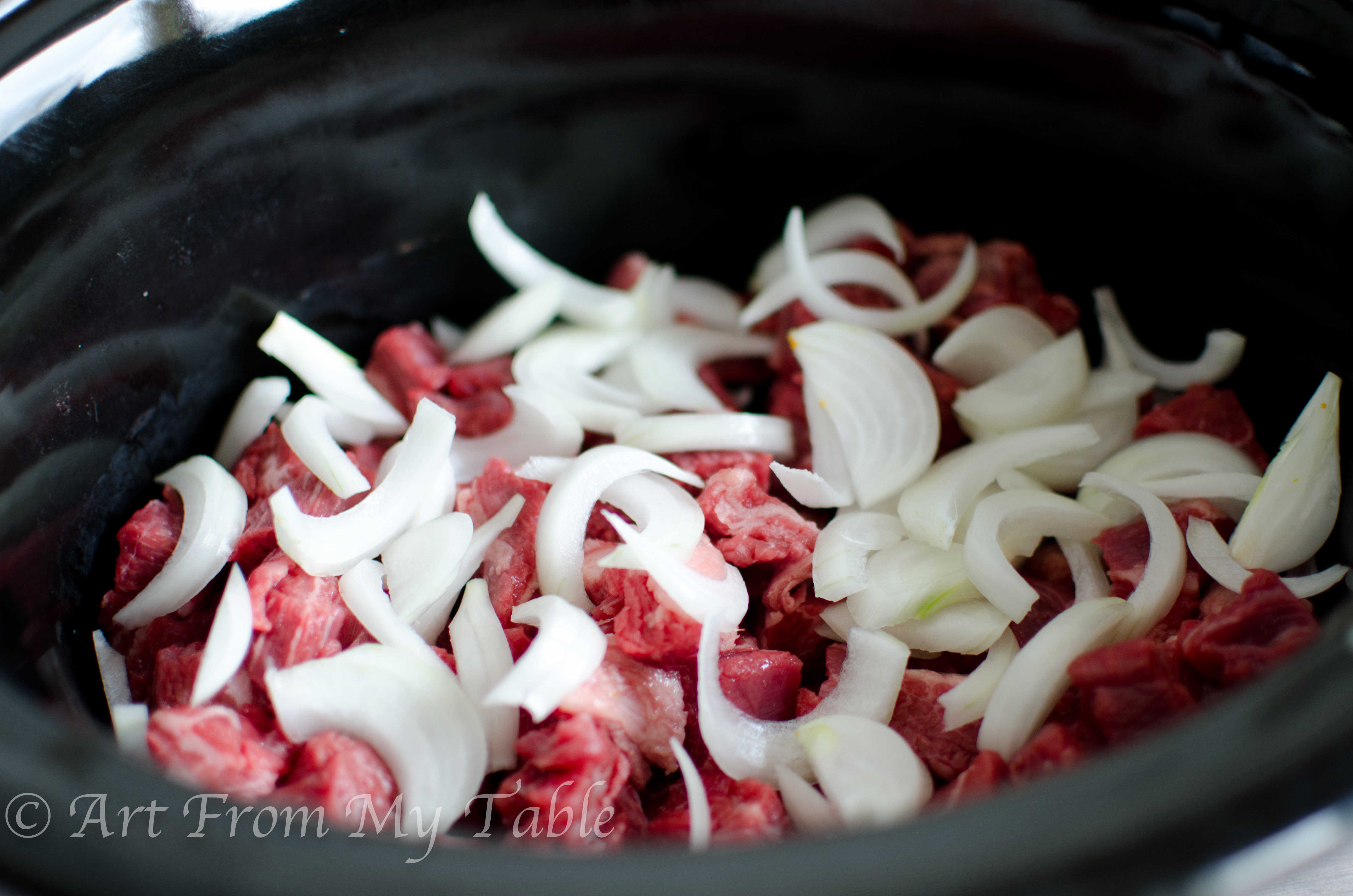 Pieces of raw steak and sliced onions in a slow cooker.