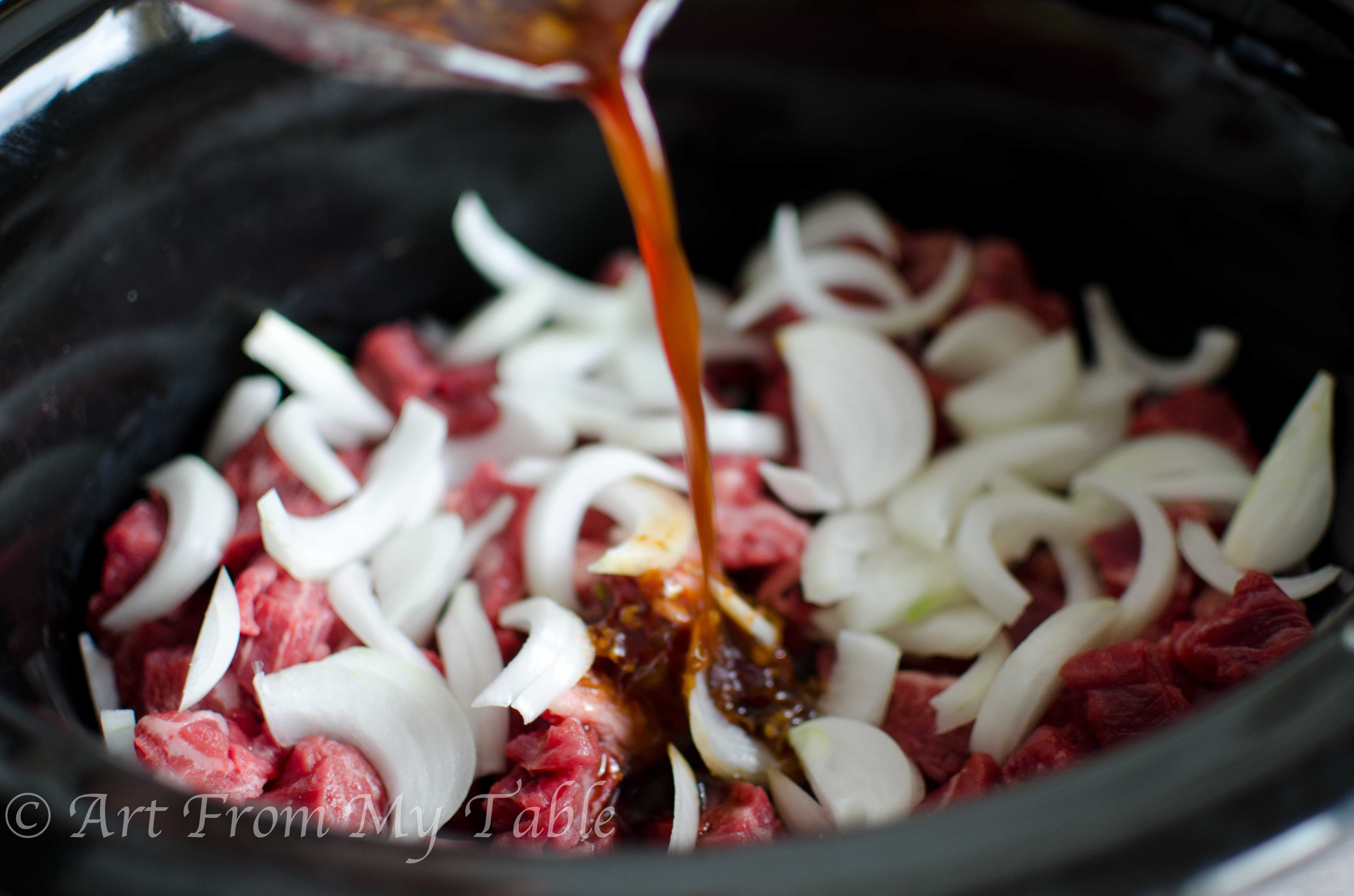 Sauce being poured over the beef and onions in a slow cooker. 