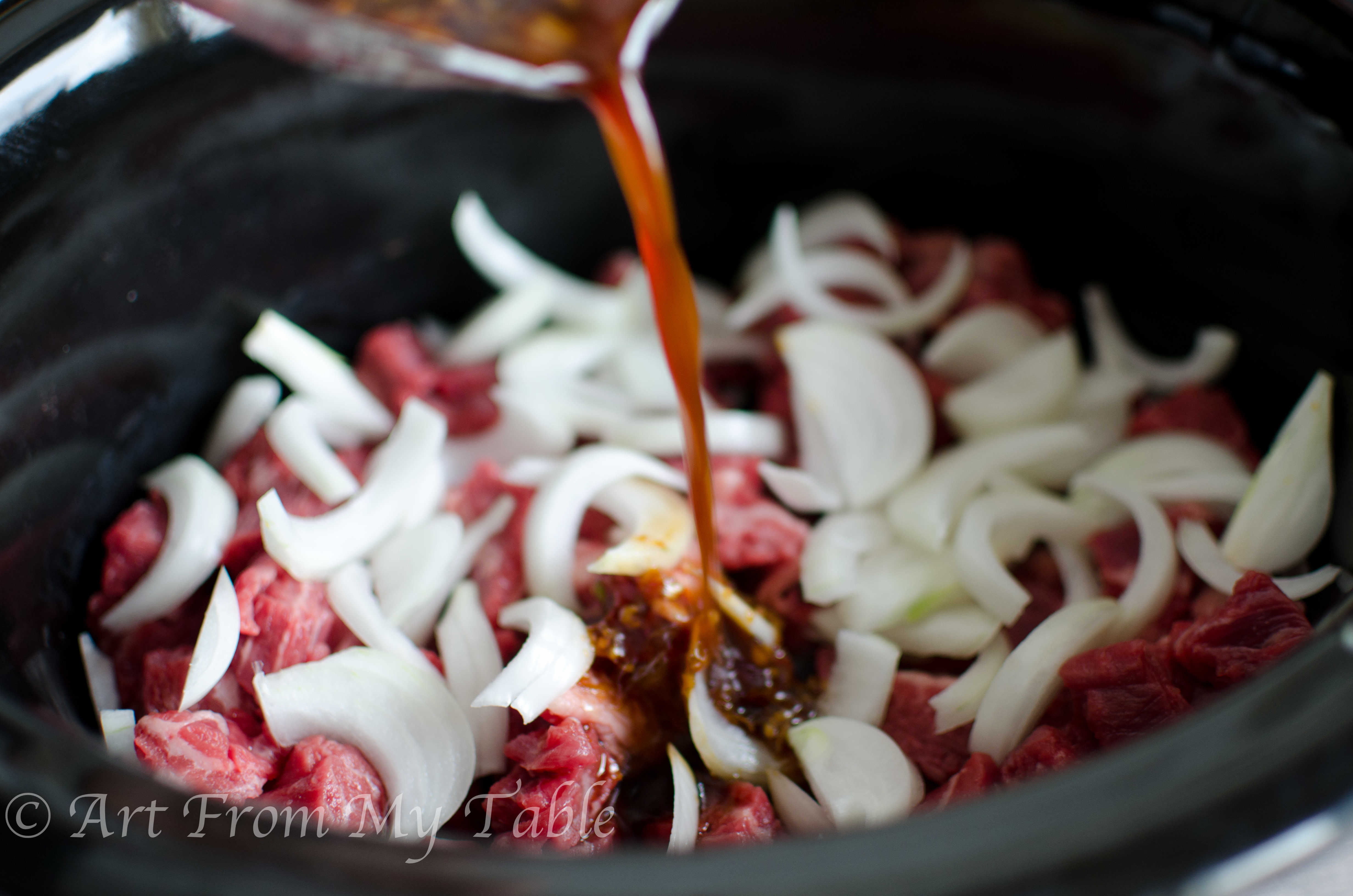 Sauce being poured over the beef and onions in a slow cooker.