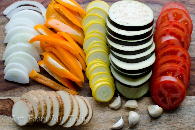 Sliced onions, orange bell pepper, yellow squash, eggplant, tomatoe, potatoes, and garlic cloves on  a cutting board.