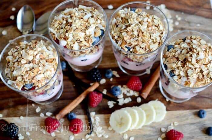 4 Banana split breakfast parfaits in a row with fresh bananas and berries surrounding them.
