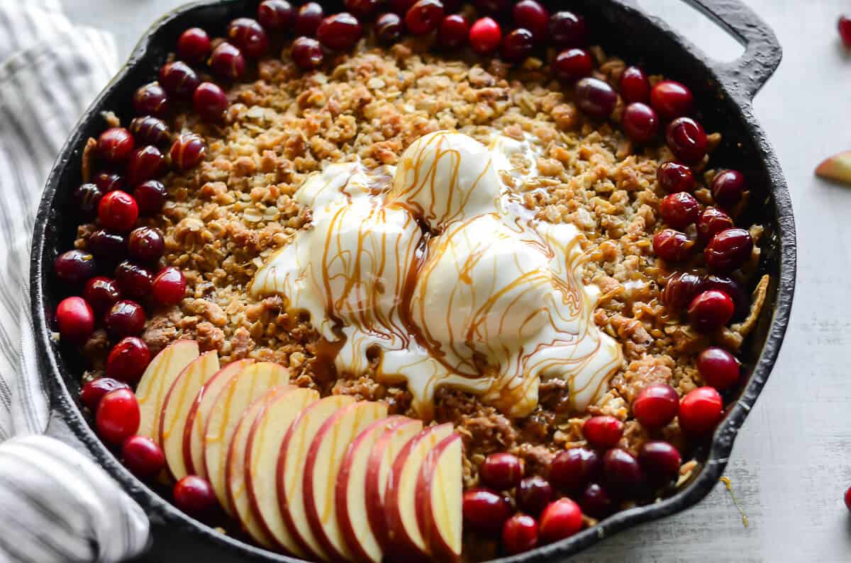 Maple apple crisp baked in a cast iron skillet topped with 3 scoops of vanilla ice cream, fresh cranberries, fresh apples and salted caramel drizzle