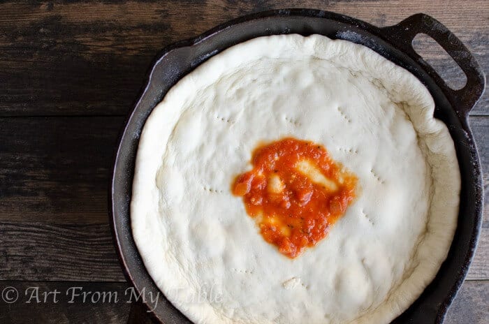 Pizza dough in a cast iron pan with a dollop of pizza sauce in the center. Dough has been pricked with a fork several times. 