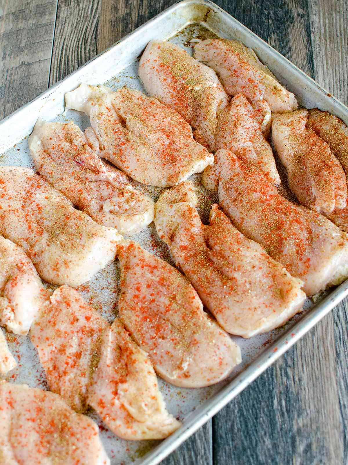 raw chicken breasts with various seasonings on a sheet pan