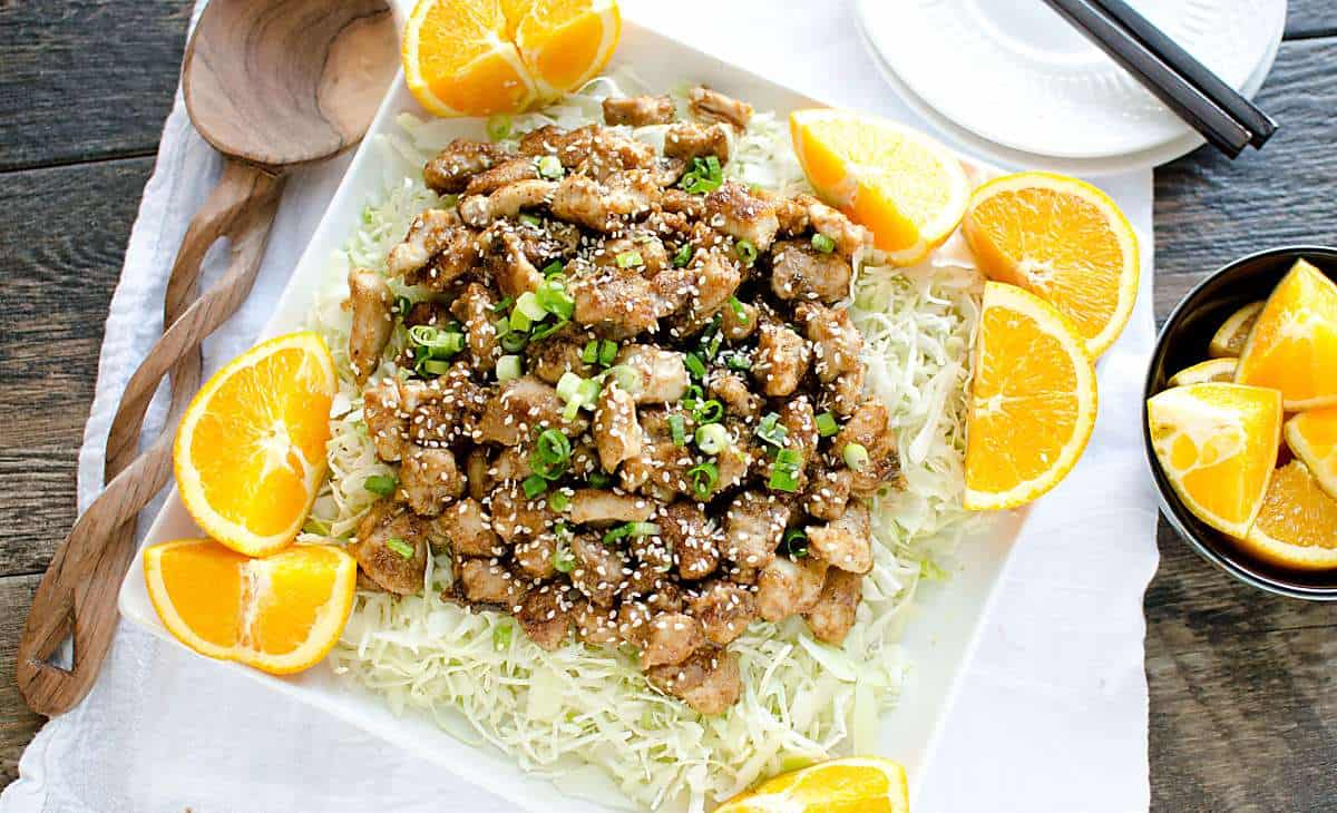 platter of homemade orange chicken on a bed of cabbage with orange slices on the side.