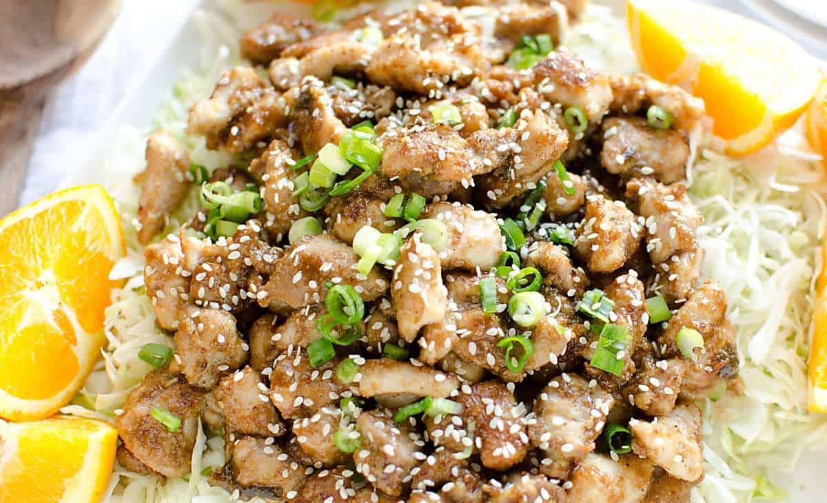 skinny orange chicken on a bed of cabbage garnished with green onions, sesame seeds and orange slices.