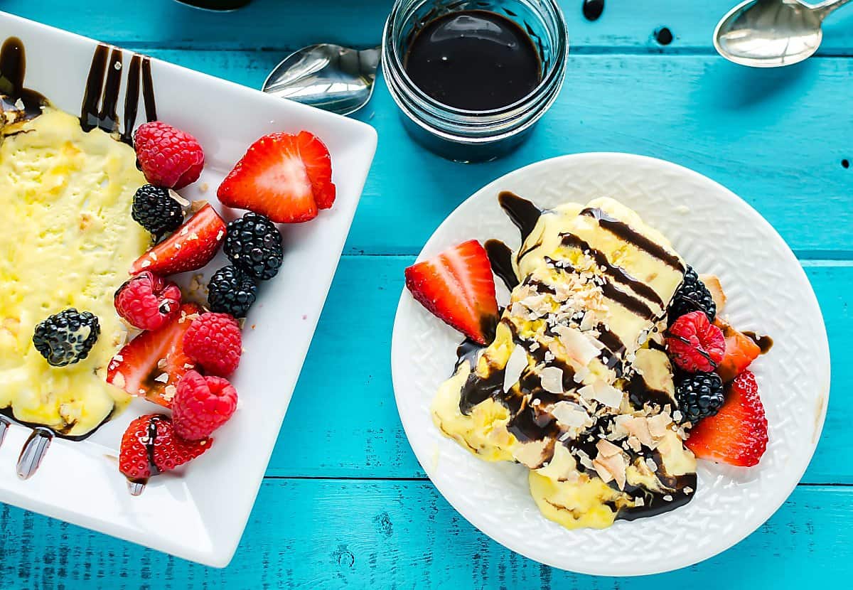 plate with 2 slices of frozen coconut parfait drizzled with chocolate syrup and garnished with strawberries, raspberries and black berries. Plate is next to platter of the traditional French parfait.