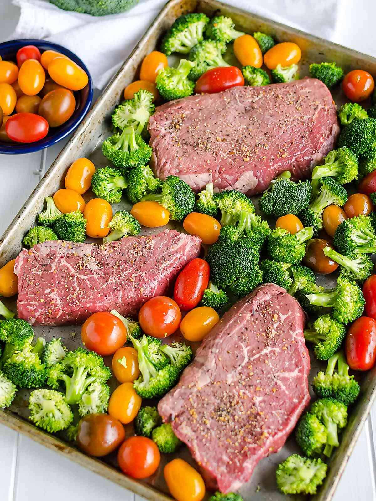 Steak, broccoli, and multi colored cherry tomatoes spread neatly on a sheet pan before cooking. Steak is seasoned with steak seasoning.