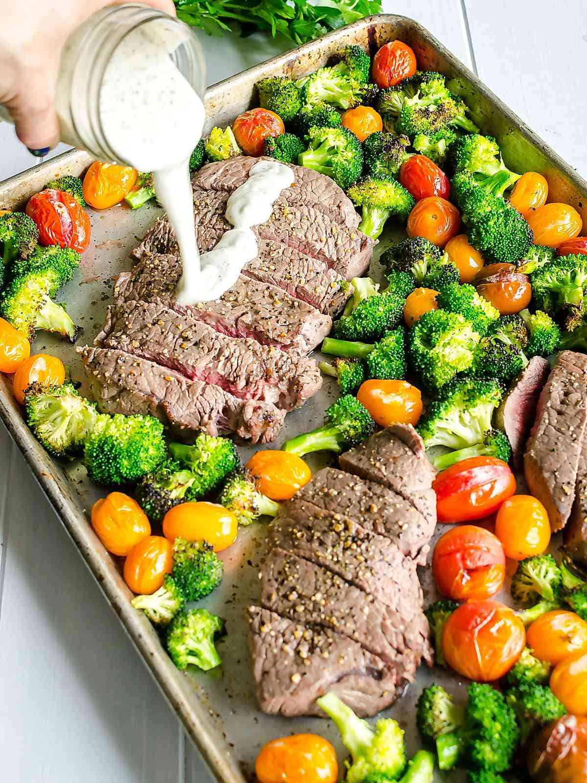 Homemade boursin cheese sauce being poured over sliced steak on a sheet pan surrounded with broccoli and cherry tomatoes.