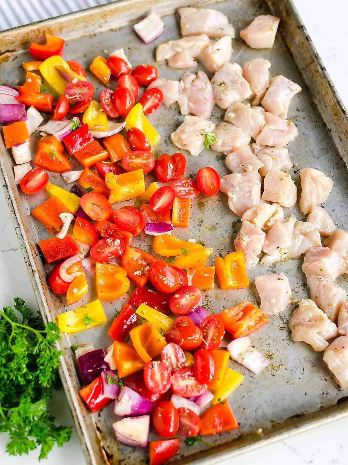 uncooked diced chicken flavored Greek style on a sheet pan with chopped colored bell peppers, tomatoes, and red onions.