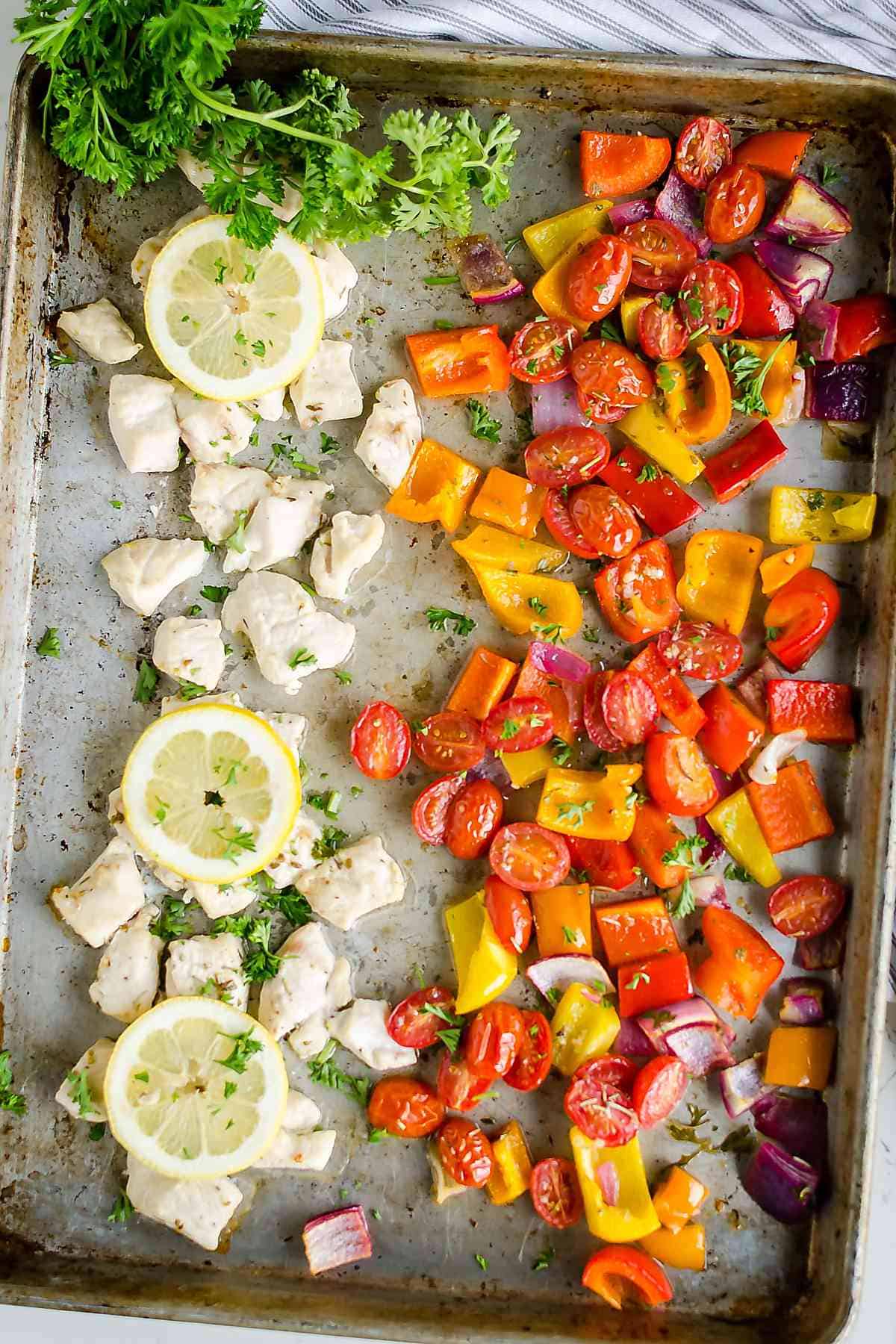 Greek style Chicken on a sheet pan with chopped colored bell peppers, grape tomatoes, red onion, lemon slices and parsley.