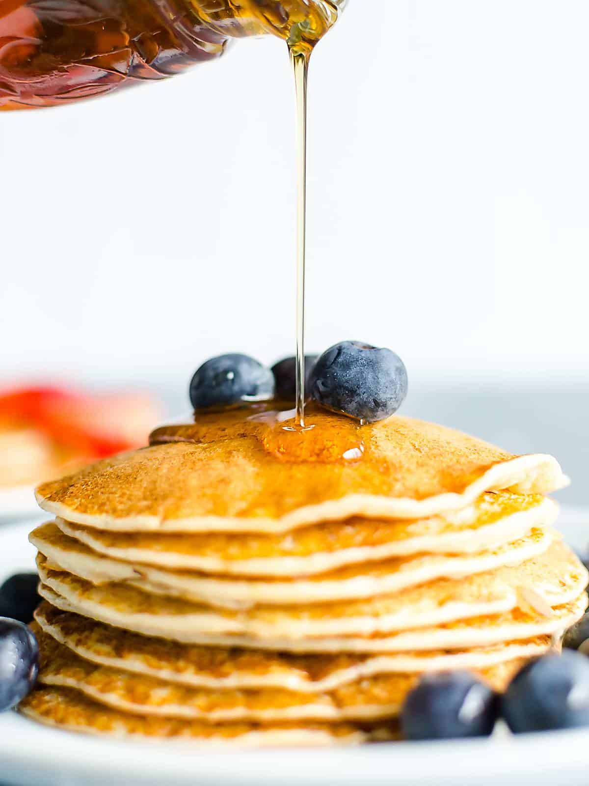 maple syrup being poured over a stack of pancakes garnished with fresh blueberries.