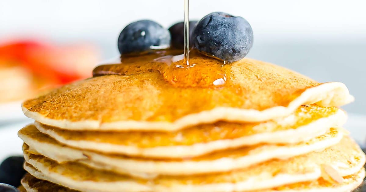 stack of lemon ricotta pancakes garnished with fresh blueberries and maple syrup being poured over the top.