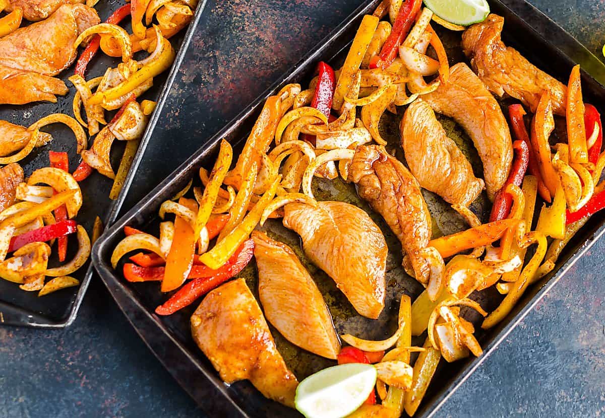 seasoned chicken tenders and sliced bell peppers and onions spread neatly on 2 sheet pans before cooking.