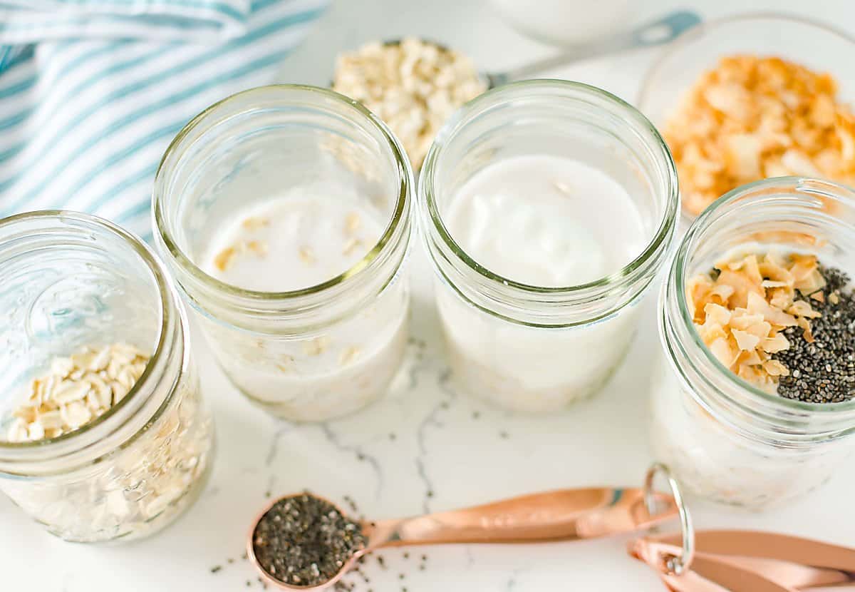4 jam jars being filled with oats, milk, yogurt, chia seeds and coconut.