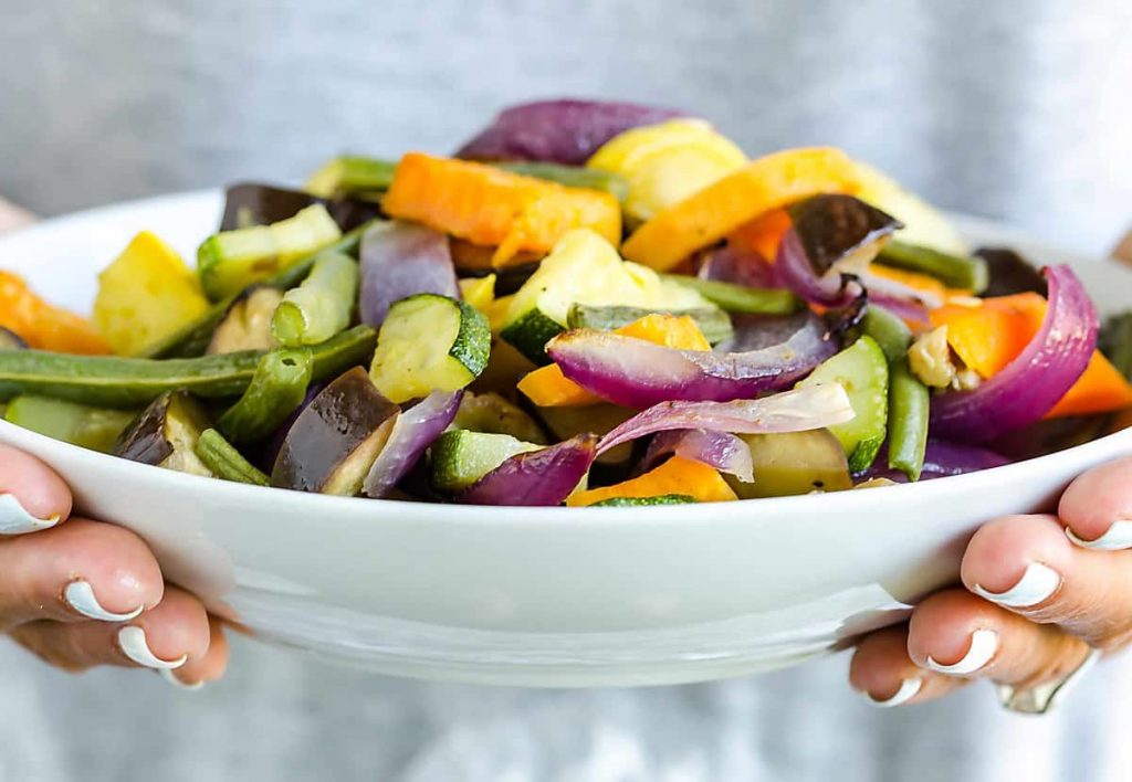 hands holding a white bowl full of colorful roasted vegetables