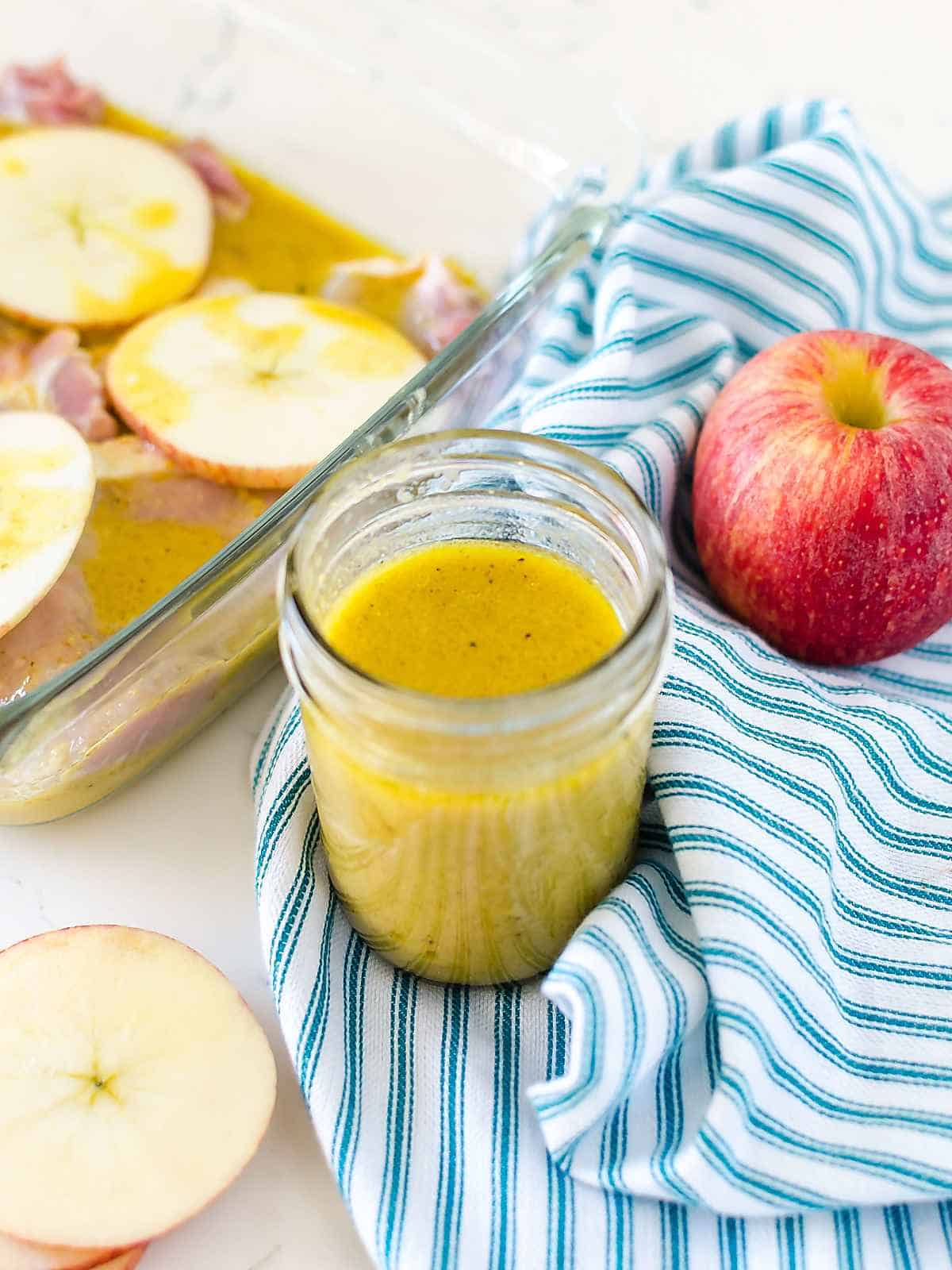small jar of apple cider mustard marinade next to fresh apple and a pan of pork chops and apples next to it in the marinade