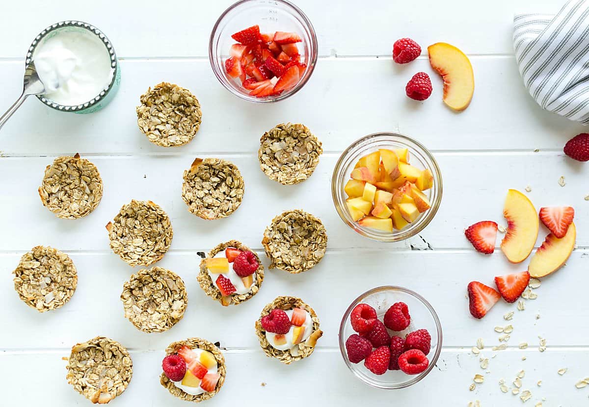 a group of coconut granola cups, some empty, some filled with yogurt, peaches, strawberries and raspberries. small bowl of yogurt on the side, small bowl of each of the fruits on the side