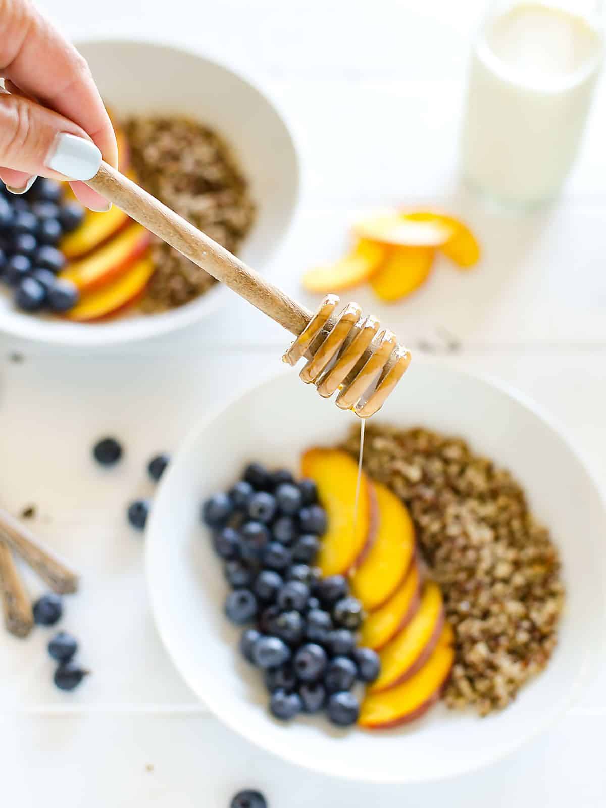 a hand holding a honey dipper over a bowl of blueberries, peaches, and quinoa, which is arranged in rows.
