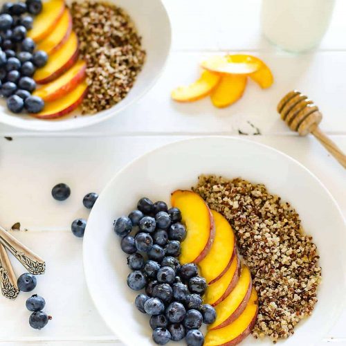two quinoa and fruit breakfast bowls, blueberries and peaches and quinoa is arranged in rows, 2 spoons off to the side, a few blueberries and peach slices on the table and a honey dipper