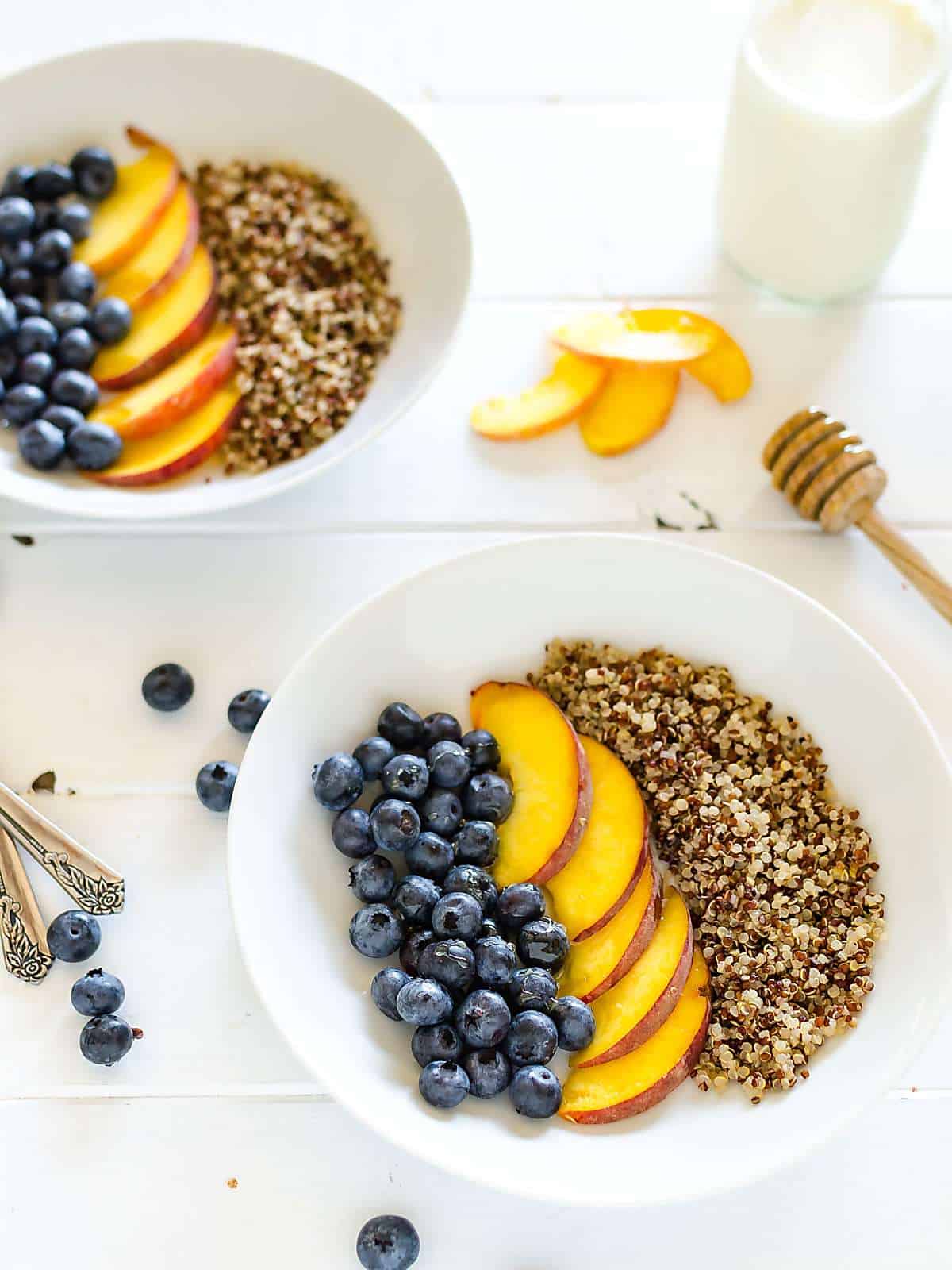 two quinoa and fruit breakfast bowls, blueberries and peaches and quinoa is arranged in rows, 2 spoons off to the side, a few blueberries and peach slices on the table and a honey dipper