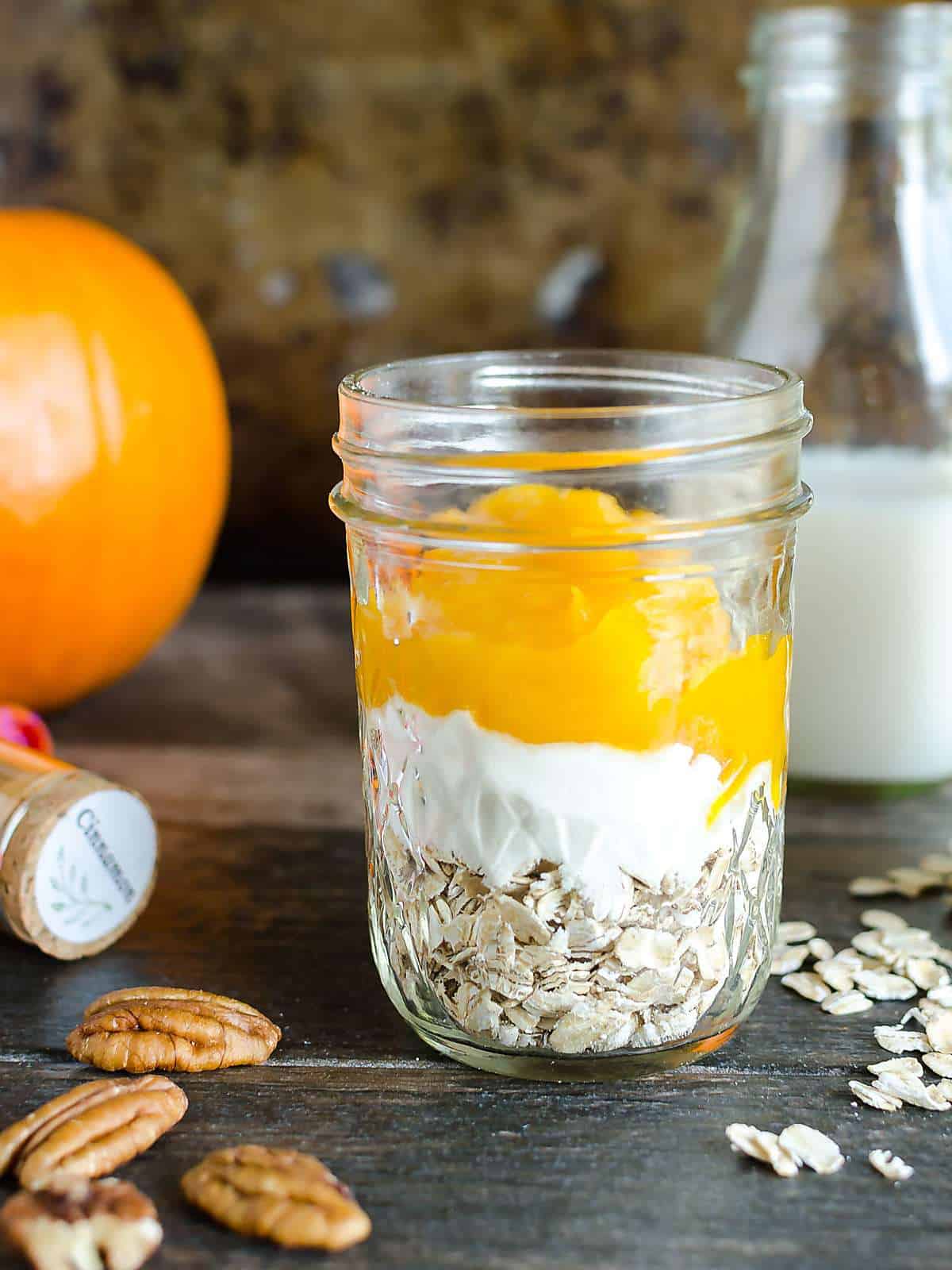 mason jar layered with rolled oats at the bottom, then a layer of protein powder, a layer of fresh pumpkin puree, jar of milk in the background, some oats spilled on the table