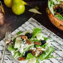 a plate full of mixed green salad recipe that contains field greens, fresh pear slices, black berries, crumbled white cheese and pecan halves, bowl of salad in the background and 2 full pears