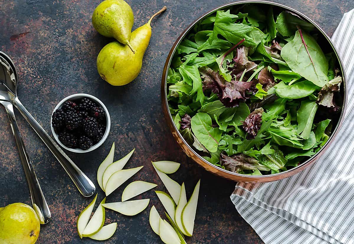 copper bowl of mixed greens, fresh pear slices on the table beside the bowl, small round container of fresh blackberries, 2 whole pears