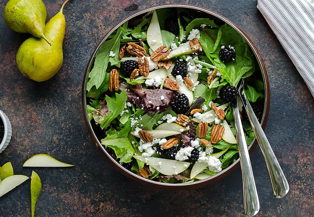 Mixed greens salad in a copper bowl full of mixed field greens, fresh pear slices, blackberries, pecans and crumbled goat cheese. 2 whole pears sitting beside it with a few pear slices