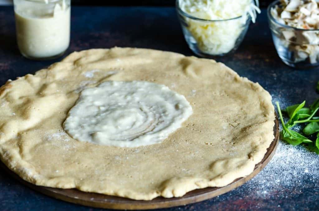 rolled out pizza crust for homemade white pizza with the start of white sauce spread in the middle, measuring cup of cheese in the background