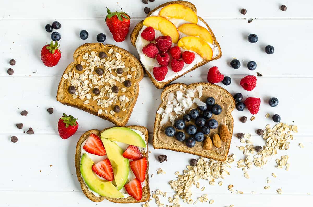 4 breakfast toast ideas, 1 slice with peanut butter, rolled oats and chocolate chips, 1 with yogurt, honey, fresh raspberries and fresh peaches, 1 with almond butter, coconut and fresh blueberries, 1 with cream cheese, avocado slices, and sliced strawberries