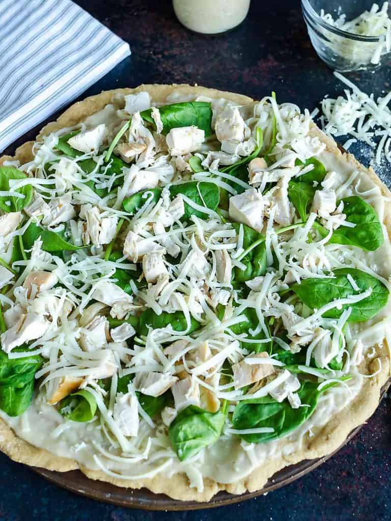 homemade white pizza ready to put into the oven topped with white sauce, chicken, spinach leaves and cheese
