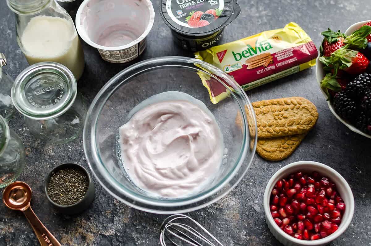 bowl of Oikos mixed berry yogurt, belVita breakfast biscuits, in the package and 2 out of the package, pomegranate arils, clear jars, and a whisk, ingredients for yogurt chia pudding
