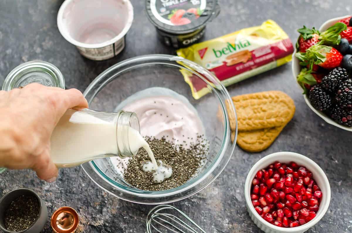 bowl of yogurt and chia seeds, milk being poured in. pomegranate arils, belVita breakfast biscuits and Oikos yogurt in the background