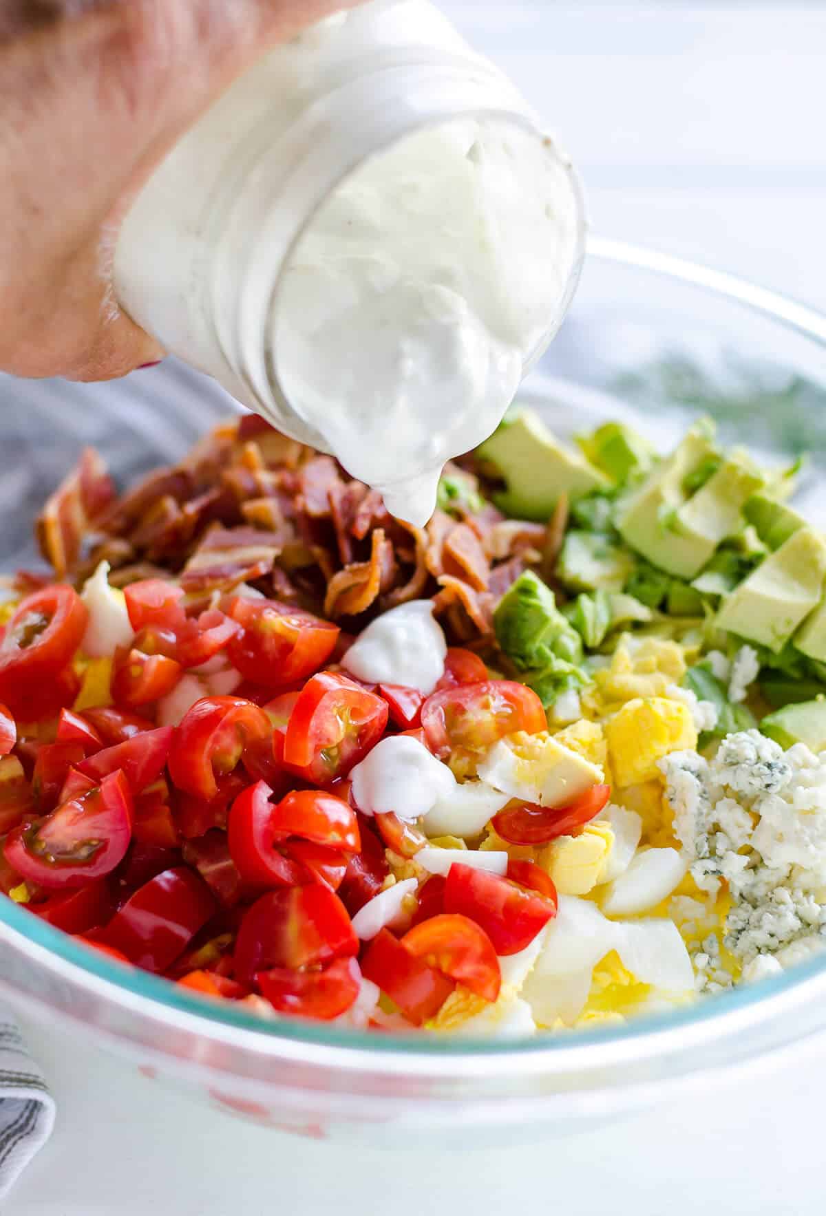 homemade blue cheese dressing being poured over chopped eggs, tomatoes, bacon, avocado and blue cheese