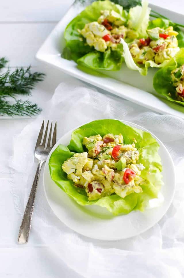 white plate with a lettuce cup filled with cobb egg salad, platter of egg salad cups in the back ground