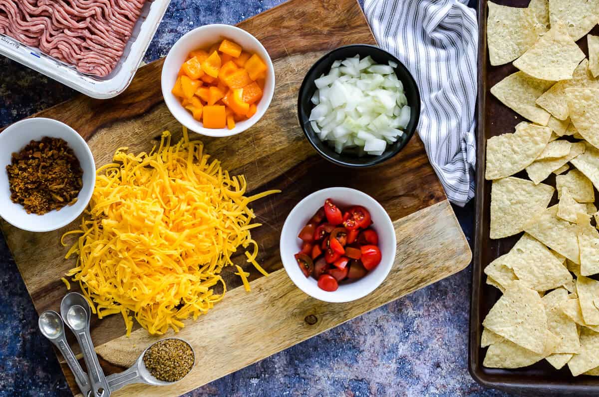 ingredients for ground turkey nachos recipe: pan of tortilla chips, shredded cheese, diced bell peppers, diced onions, diced tomatoes and raw ground turkey