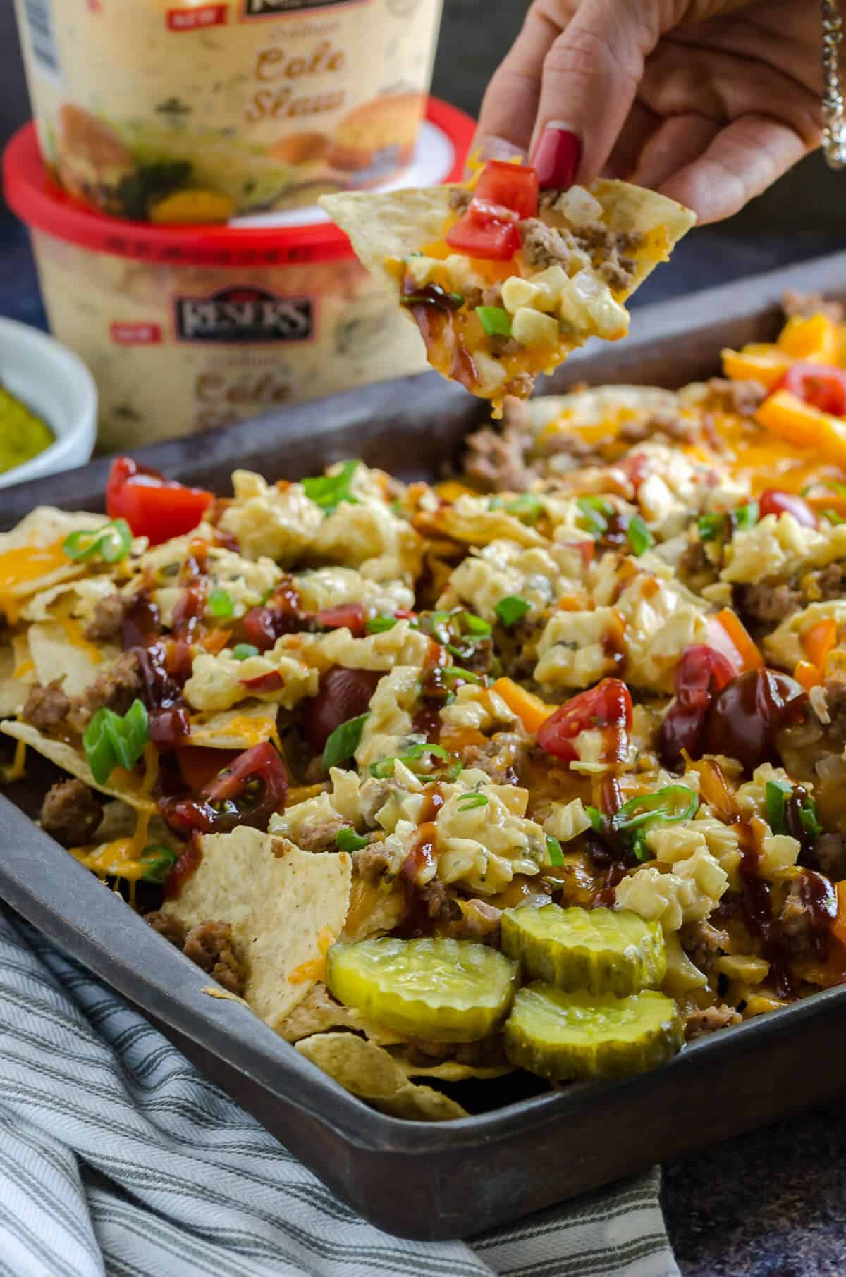 hand pulling a chip off a pan of ground turkey nachos topped with coleslaw, barbecue sauce and a side of pickles. Resers stadium cole slaw in the background