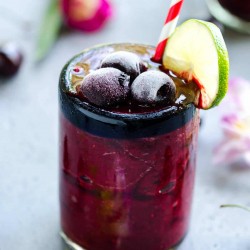 glass of deep red frozen cherry cooler garnished with sweet cherries and a lime slice. flowers in the back ground