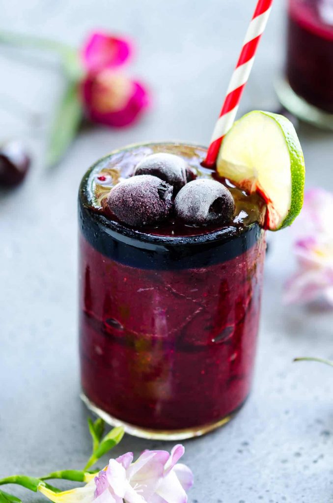 glass of deep red frozen cherry cooler garnished with sweet cherries and a lime slice. flowers in the back ground