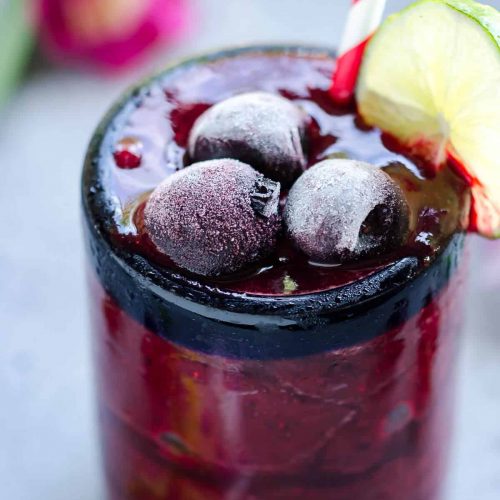 deep red frozen cherry cooler filled to the brim of a clear glass topped with frosty frozen sweet cherries and garnished with slice of lime on the side of the glass