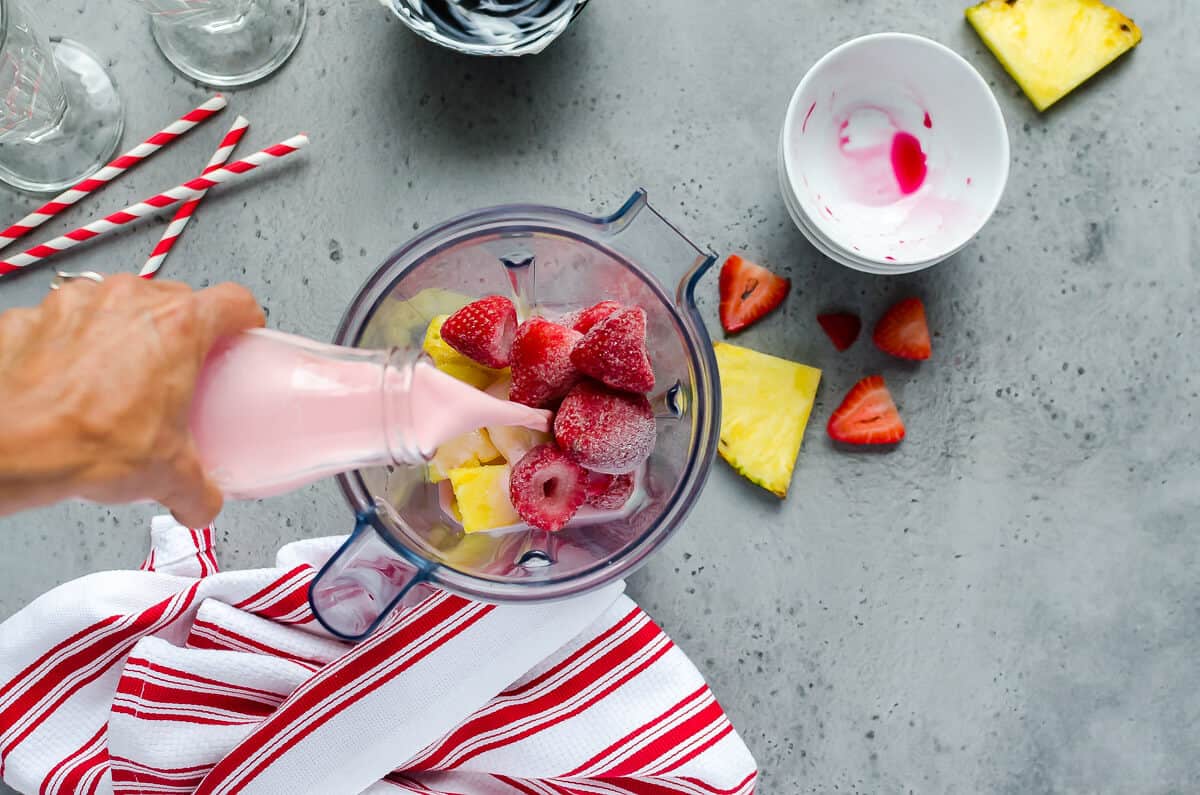 hand pouring strawberry milk into a blender filled with frozen strawberries and pineapples