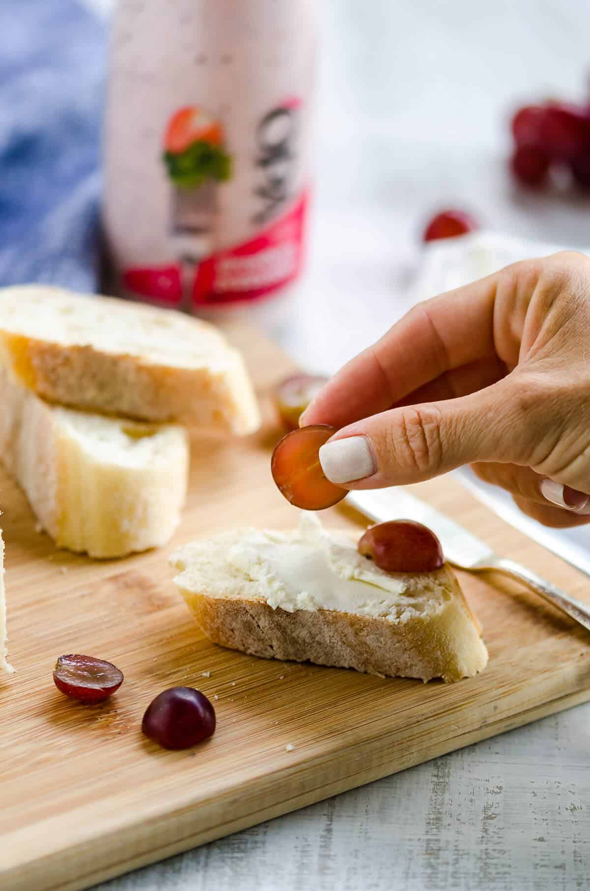 crostini with goat cheese on a cutting board, hand placing grapes on it.